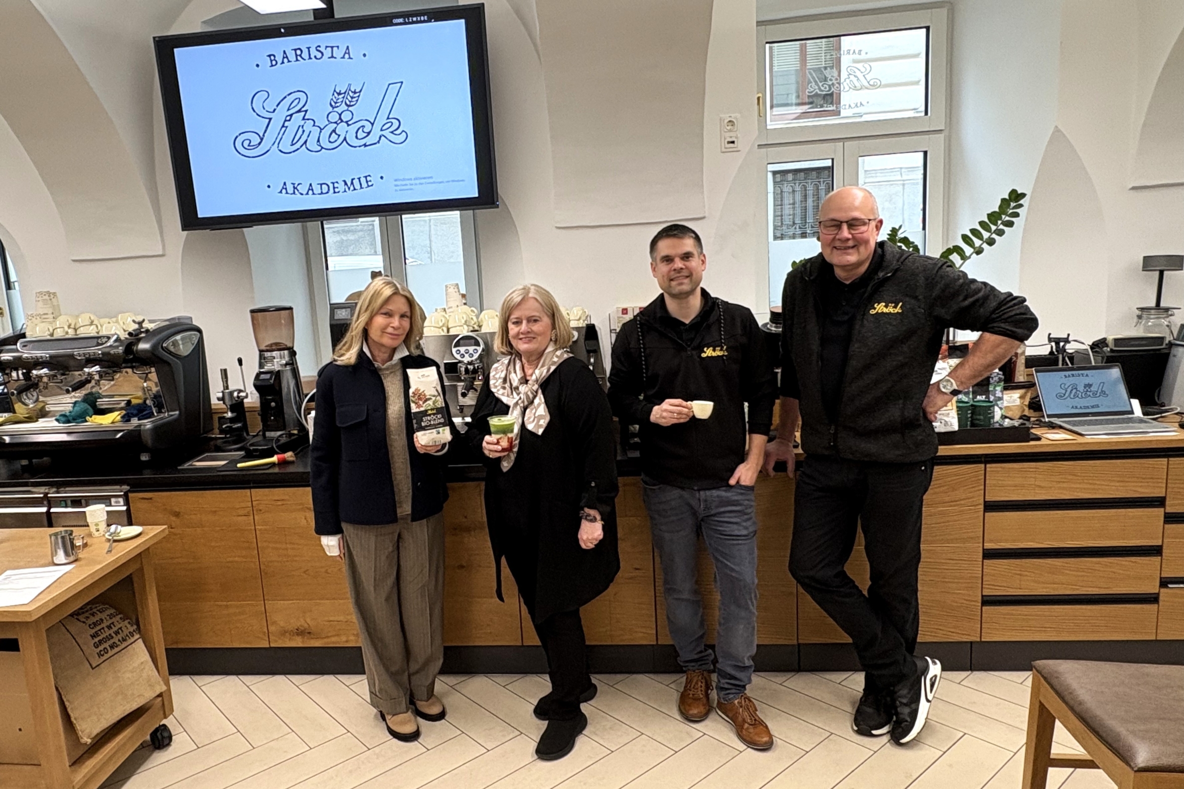 Vier Personen stehen in einer modernen Kaffee-Akademie und lächeln in die Kamera. Hinter ihnen befindet sich eine Theke mit Kaffeegeräten und einem Bildschirm, auf dem die Barista Ströck Akademie ihre Spezialität Bio-Eis-Ichigo-Matcha anpreist.