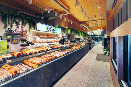 Eine moderne Bäckerei mit einer langen Auslage, die mit verschiedenen Gebäcken, Broten und Backwaren gefüllt ist. Der Laden ist hell beleuchtet, die Speisekarten sind digital und grüne Pflanzen schmücken die Wände und die Decke.