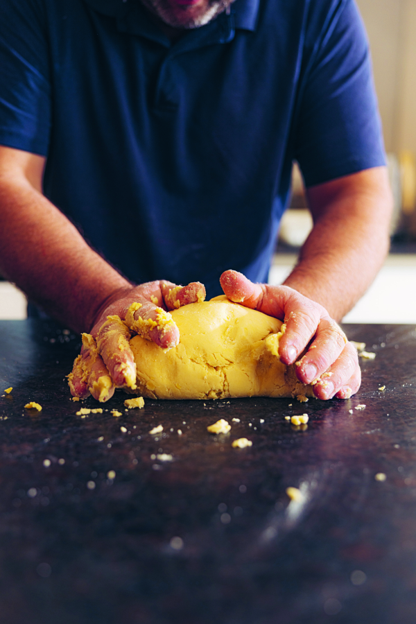 Eine Person knetet gelben Teig auf einer dunklen Arbeitsplatte und trägt ein marineblaues Hemd.