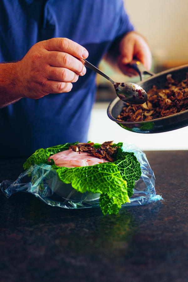 Eine Person löffelt gekochte Pilze aus einer Pfanne auf Schinkenscheiben und Wirsingblätter, die auf Frischhaltefolie auf einem Küchentisch liegen.