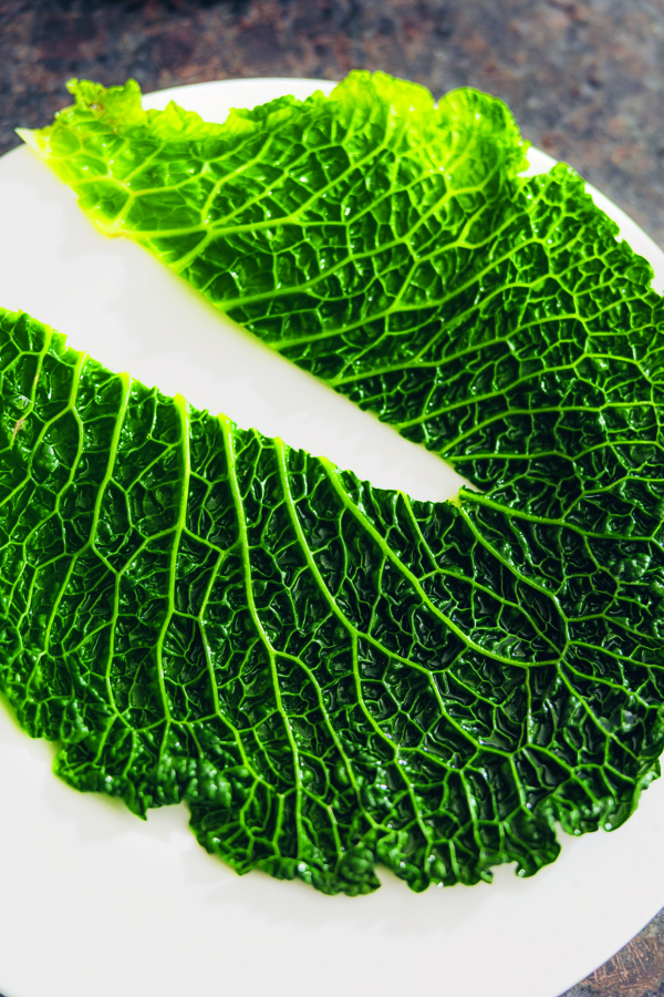 Zwei große, leuchtend grüne Wirsingblätter sind auf einem weißen Teller angeordnet und zeigen ihre strukturierte, zerknitterte Oberfläche und ihre ausgeprägten Adern. Der Hintergrund ist dunkel und unscharf.