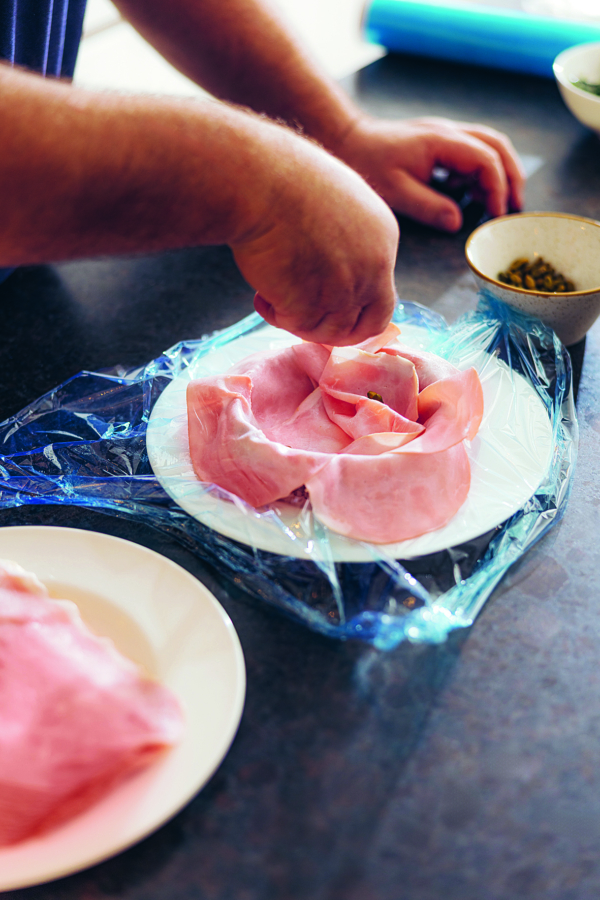 Eine Person ordnet Schinkenscheiben kreisförmig auf einem weißen, mit Plastikfolie bedeckten Teller an, während Schüsseln und zusätzlicher Schinken in der Nähe auf einer dunklen Arbeitsplatte stehen.