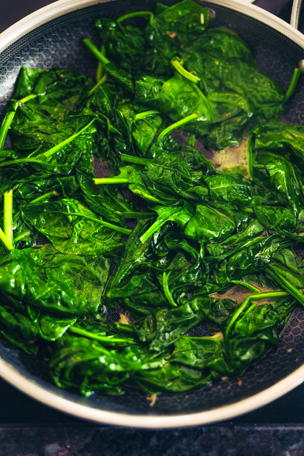 Leuchtend grüne Spinatblätter werden in einer Pfanne gebraten, wobei einige von der Hitze und der Feuchtigkeit verwelkt sind und glänzen, wodurch ein frisches und gesundes Gericht entsteht.
