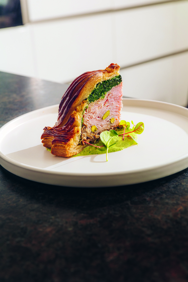 Eine Scheibe Beef Wellington mit goldener Blätterteigkruste, rosa Rindfleisch in der Mitte, grünem Spinat und garniert mit Mikrogrün, serviert auf einem weißen Teller auf einer dunklen Arbeitsplatte.