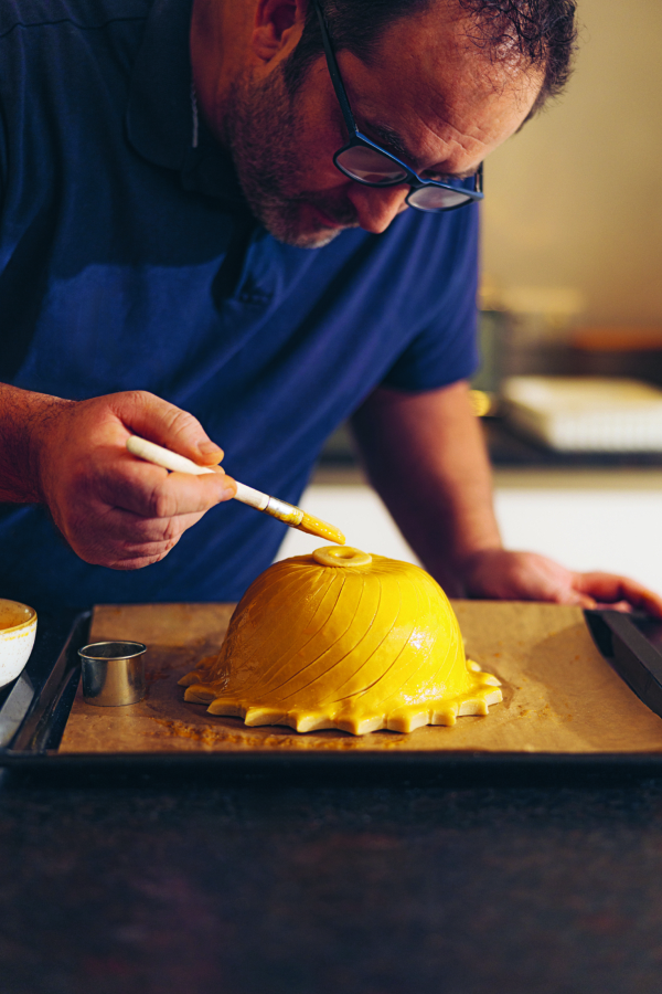 Eine Person, die eine Brille und ein marineblaues Hemd trägt, pinselt in einer Küche vorsichtig eine Eimasse auf eine runde, dekorative Gebäckkuppel auf einem Backblech.