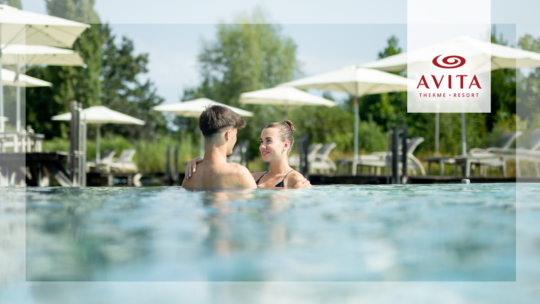 Ein Paar steht eng beieinander in einem Freibad und lächelt sich gegenseitig an. Im Hintergrund sind Liegestühle und Sonnenschirme sowie Bäume zu sehen. Das Logo des AVITA Therme Resorts befindet sich auf der rechten Seite des Bildes.