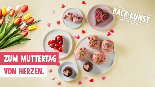 Verschiedene herzförmige und runde Gebäckstücke, einige mit rosa Zuckerguss oder Erdbeeren, sind auf Tellern neben einem Tulpenstrauß angeordnet. Der Text lautet: Zum Muttertag von Herzen. BACK KUNST seit 1970.