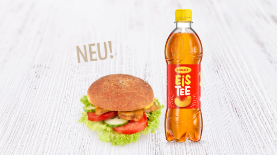 Ein Sandwich mit Salat, Tomate, Gurke und einer panierten Füllung steht neben einer Flasche Rauch Eistee Pfirsich-Eistee auf einer weißen Holzfläche. Im Hintergrund ist das Wort "NEU!" zu lesen.