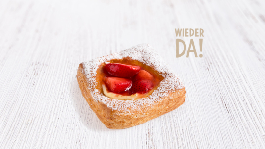 Ein quadratischer, mit Erdbeeren belegter und mit Puderzucker bestäubter Kuchen liegt auf einer weißen Holzfläche. Der Schriftzug Wieder da! erscheint in hellbrauner Schrift im Hintergrund.