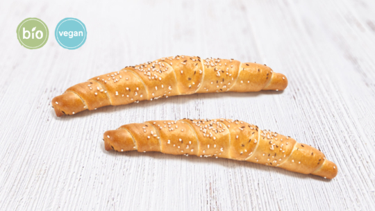 Zwei halbmondförmige Brötchen mit Sesam auf der Oberseite, die auf einer weißen Holzunterlage liegen. Auf den kreisförmigen Etiketten in der Ecke steht "bio" und "vegan", was bedeutet, dass die Brötchen biologisch und vegan sind.