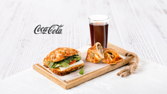 Ein Holztablett mit einem Sandwich, zwei Gebäckstücken und einem Glas dunkler Limonade. Der Hintergrund ist weiß, und das Coca-Cola-Logo ist sichtbar.