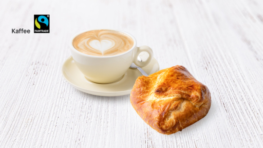 Eine Tasse Cappuccino mit herzförmigem Milchkaffee steht auf einer Untertasse mit Löffel, daneben ein goldbraunes Gebäck auf einer weißen Holzfläche. Das Fairtrade-Logo und das Wort Kaffee sind in der oberen linken Ecke zu sehen.