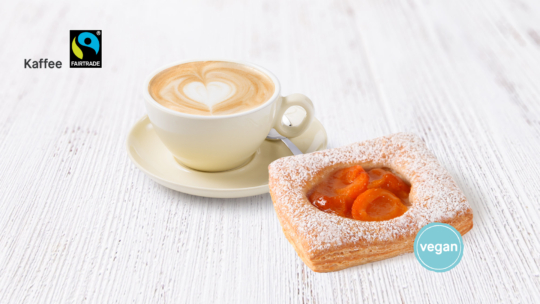 Eine Tasse Cappuccino mit herzförmigem Schaum sitzt auf einer Untertasse neben einem veganen, mit Puderzucker bestäubten Aprikosenkuchen. Fairtrade- und vegane Etiketten befinden sich auf einer weißen Holzoberfläche.