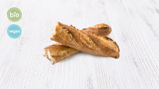 Zwei knusprige Baguettes lagen auf einem weißen Holzhintergrund übereinander. Auf den runden Aufklebern in der oberen linken Ecke stand „bío“ und „vegan“.