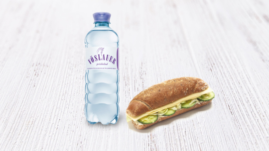 Eine Flasche Vöslauer Mineralwasser und ein Sandwich mit Käse, Salat und Gurke auf einer Holzunterlage.