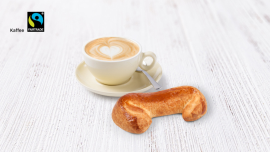 Auf einem hellen Holztisch steht eine Tasse Cappuccino mit einem Herzmuster im Schaum auf einer Untertasse, daneben liegt ein halbmondförmiges Gebäckstück mit einem Löffel. In der oberen linken Ecke ist das Fairtrade-Logo zu sehen.