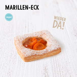 Auf einer weißen Holzunterlage liegt ein quadratisches, mit Puderzucker bestreutes Aprikosen-Plundergebäck. Der Text lautet „MARILLEN-ECK“, „vegan“ und „WIEDER DA!“, was darauf hinweist, dass das Gebäck vegan ist und wieder im Sortiment ist.