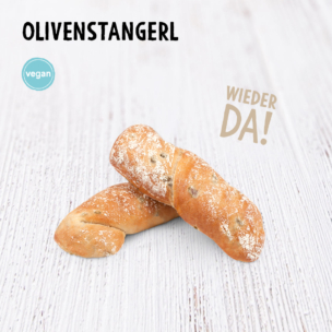 Zwei mit Mehl bestäubte Olivenbrotstangen liegen auf einer hellen Holzunterlage. Der Text darüber lautet „OLIVENSTANGERL“, mit den Bezeichnungen „vegan“ und „WIEDER DA!“ auf Deutsch, was so viel bedeutet wie „Olivenstangen“ und „wieder da“.