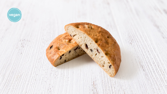 Ein rundes Brot mit Oliven wird in zwei Hälften geschnitten und auf eine weiße Holzunterlage gelegt. In der oberen linken Ecke ist ein blaues Vegan-Label zu sehen.