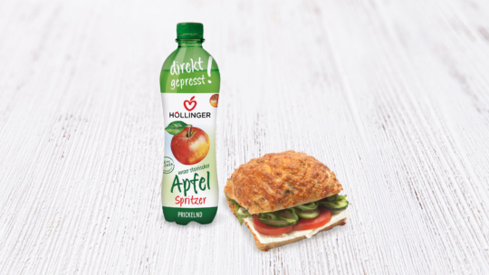 Auf einer weißen Holzplatte steht eine Flasche Höllinger Apfelspritzer neben einem Sandwich mit Gemüse und Käse auf einem Blätterteigbrötchen.