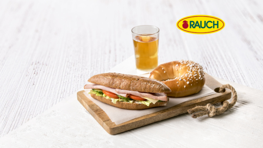 Ein Sandwich mit Schinken, Salat und Tomate, ein gesalzenes Brezelbrötchen und ein Glas Eistee auf einem Holzbrett. Das RAUCH-Logo ist in der oberen rechten Ecke auf weißem Hintergrund zu sehen.