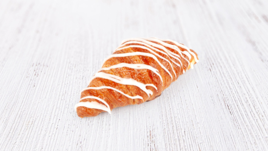 Ein goldbraunes Croissant mit weißer Glasur darüber, das auf einer hellen Holzunterlage liegt.
