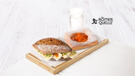 Ein Sandwich mit Käse und Salat, ein mit Puderzucker bestäubtes Aprikosengebäck und ein Glas Milch auf einem Tablett. In der oberen rechten Ecke ist das Römerquelle-Logo zu sehen.