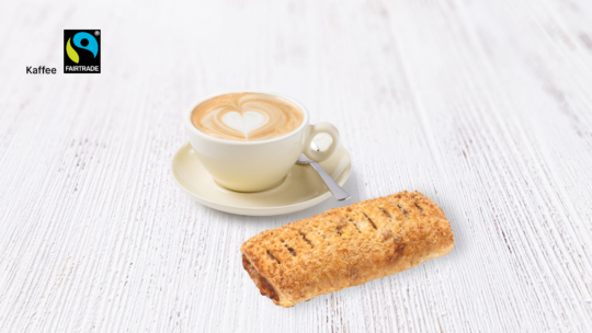Auf einer weißen Holzplatte steht eine Tasse Latte mit herzförmigem Milchschaum neben einem Gebäckstück auf einer Untertasse. In der oberen linken Ecke ist ein Fairtrade-Kaffee-Logo zu sehen.