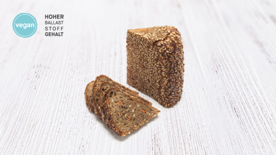 Ein Laib Vollkornbrot mit Körnern, teilweise in Scheiben geschnitten, liegt auf einer hellen Holzunterlage. In der oberen linken Ecke sind ein blaues Vegan-Label und der deutsche Text „HOHER BALLASTSTOFFGEHALT“ zu sehen.