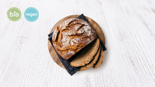 Ein rundes Schwarzbrot mit drei Scheiben auf einem Holzbrett, darunter eine schwarze Serviette, und Bio- und Vegan-Siegel in der oberen linken Ecke auf einer weißen Holzfläche.