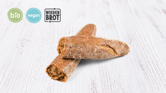 Zwei Scheiben Vollkornbaguette auf einer weißen Holzunterlage, darüber Etiketten mit den Aufschriften „Bio“, „vegan“ und „Wieder Brot“.