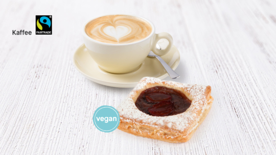 Auf einer weißen Holzplatte steht eine Tasse Cappuccino mit herzförmiger Latte-Art neben einem veganen Gebäckstück, das mit Marmelade und Puderzucker verziert ist. Fairtrade- und Vegan-Siegel sind zu sehen.