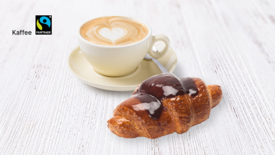 Eine Tasse Cappuccino mit herzförmiger Latte-Art steht auf einer Untertasse mit einem Löffel neben einem mit Schokolade überzogenen Croissant auf einem weißen Holztisch. In der oberen linken Ecke sind das Fairtrade- und das Kaffee-Logo zu sehen.