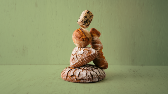 Fünf verschiedene handwerklich hergestellte Brotsorten, darunter ein rundes Brot mit Rissen, ein herzförmiges Brot mit Mandeln und Brötchen, sind kunstvoll vor einem hellgrünen Holzhintergrund gestapelt.