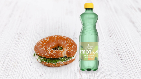 Auf einer weißen Holzplatte liegt ein Sesambagel mit Frischkäse und Kräutern neben einer grünen Plastikflasche mit „Römerquelle Emotion“-Wasser mit Birnen-Zitronenmelisse-Geschmack.