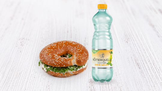 Auf einer weißen Holzplatte liegt ein Sesam-Bagel mit Frischkäse und Kräutern neben einer Flasche Römerquelle Emotion Mineralwasser mit Birnen-Melonen-Geschmack.