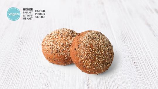 Zwei runde, mit Körnern versetzte Brötchen auf einer weißen Holzunterlage. Die Symbole auf der linken Seite weisen darauf hin, dass die Brötchen vegan sowie ballaststoff- und eiweißreich sind; daneben steht ein deutscher Text.