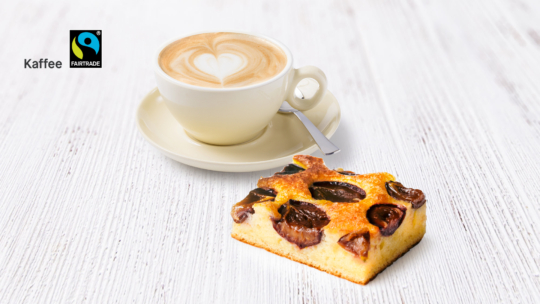 Eine Tasse Kaffee mit Latte-Art steht auf einer Untertasse mit einem Löffel daneben, neben einem Stück Kuchen mit Obstbelag auf einer weißen Holzunterlage. In der oberen linken Ecke sind das Fairtrade-Logo und das Wort „Kaffee“ zu sehen.