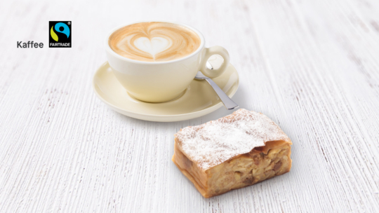Eine Tasse Cappuccino mit herzförmiger Latte-Art steht auf einer Untertasse mit einem Löffel, daneben liegt ein Stück mit Puderzucker bestreuter Apfelstrudel auf einer weißen Holzunterlage. In der oberen linken Ecke sind die Fairtrade- und Kaffee-Logos zu sehen.
