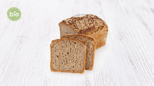 Ein Mehrkornbrot mit Sesamkörnern liegt auf einer weißen Holzunterlage. Vor dem Brot sind drei Scheiben abgeschnitten. In der oberen linken Ecke befindet sich ein grünes Bio-Siegel.