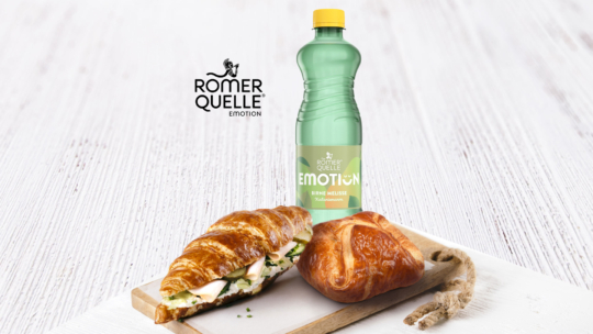 Eine grüne „Römerquelle Emotion“-Flasche steht auf einer weißen Holzunterlage neben einem Croissant-Sandwich mit Salat und Käse sowie einem einfachen Croissant auf einem Holzbrett mit Seilgriff.