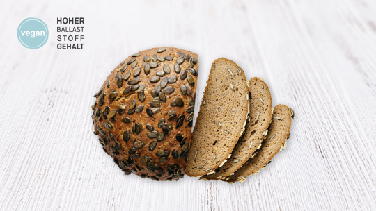 Ein Laib Brot mit Körnerkruste, aus dem drei Scheiben herausgeschnitten sind, daneben ein veganes Label und der deutsche Text HOHER BALLASTSTOFFGEHALT.