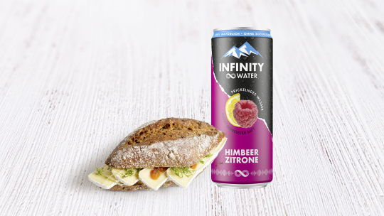 Neben einer Dose Infinity Water – Sprudelwasser mit Himbeer-Zitronen-Geschmack – liegt ein knuspriges Brötchen, gefüllt mit Brie und Kräutern.