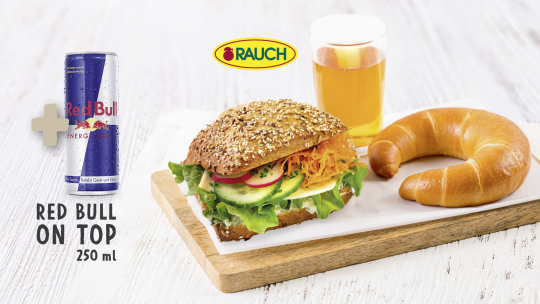 Eine Red-Bull-Dose, ein Gemüsesandwich auf einem Holzbrett, ein Glas Saft und ein Croissant mit dem Rauch-Logo darüber.