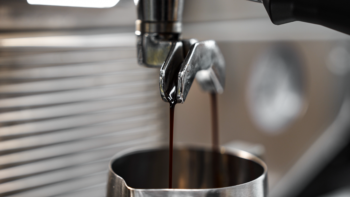 Nahaufnahme von dunklem Espresso, der aus dem Auslauf eines Portafilters einer Espressomaschine aus Metall in einen Edelstahlkrug gegossen wird, mit unscharfem Hintergrund.