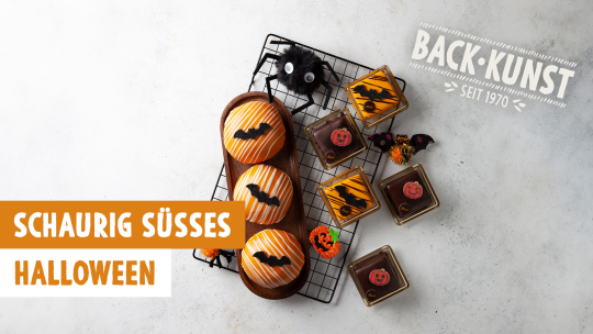Eine Auswahl an Gebäck und Kuchen zum Thema Halloween auf einem Abkühlgitter, mit den Aufschriften BACK-KUNST SEIT 1970 und SCHAURIG SÜSSES HALLOWEEN.