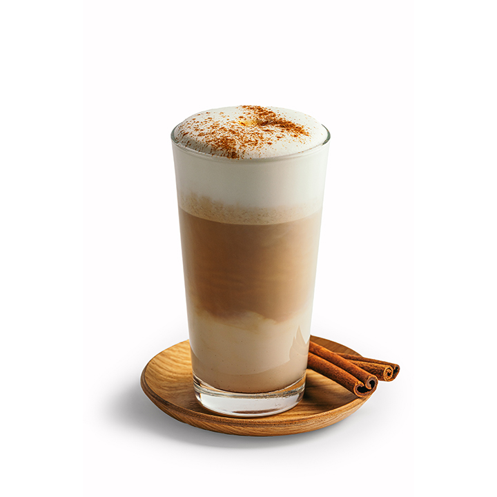 Ein Glas mit geschichtetem Kaffee, gekrönt von Milchschaum und Zimt, serviert auf einer hölzernen Untertasse mit zwei Zimtstangen.