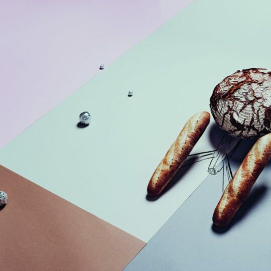 Ein runder Laib knusprigen Brotes mit Rissmustern ist über drei Baguettes positioniert, die einer Spinne ähneln. Das Brotarrangement steht auf buntem, geometrischem Papier mit verstreuten Kristallen.