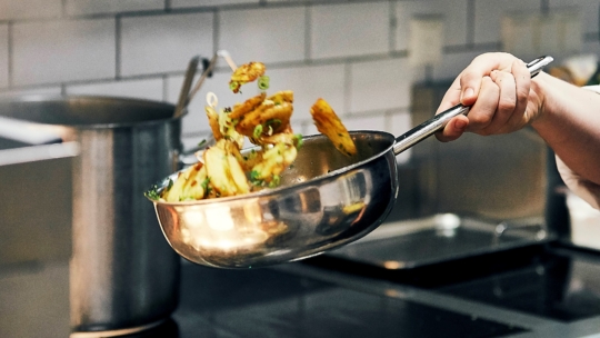 Eine Person schwenkt geschnittenes Gemüse in einer Edelstahlpfanne über einer Herdplatte in einer modernen Küche mit weißen Kachelwänden.