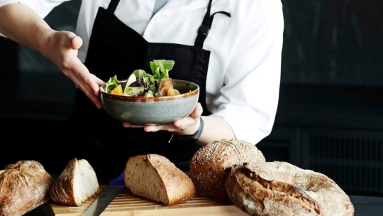 Eine Person in weißem Hemd und schwarzer Schürze präsentiert eine Schüssel mit Salat, während im Vordergrund auf einem Holzbrett mehrere Brote und rustikale Brotscheiben liegen.